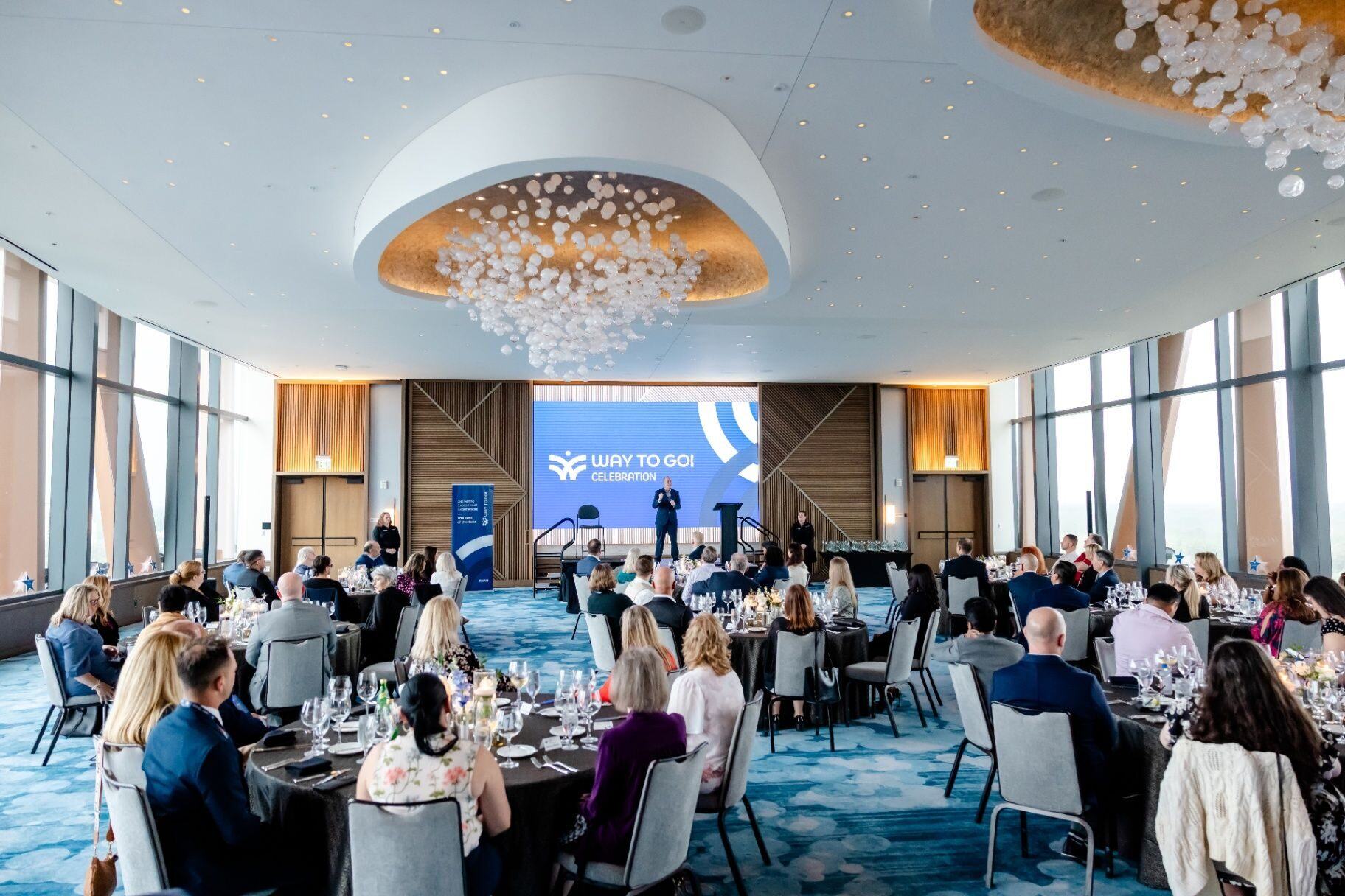 Corporate celebration event with guests seated at dinner while a speaker presents on stage